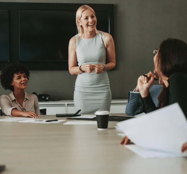 A woman in a light dress stands smiling at a conference table, engaging with colleagues. The setting is professional and collaborative, conveying positivity.