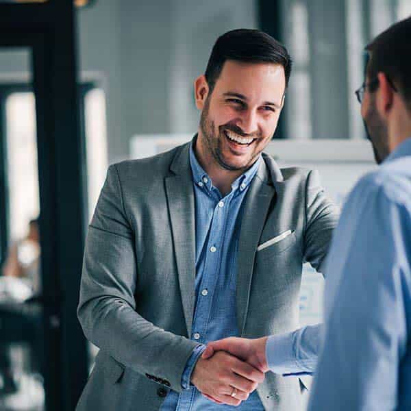 A man in a gray suit is smiling warmly as he shakes hands with another person in an office setting, conveying a sense of success and teamwork.
